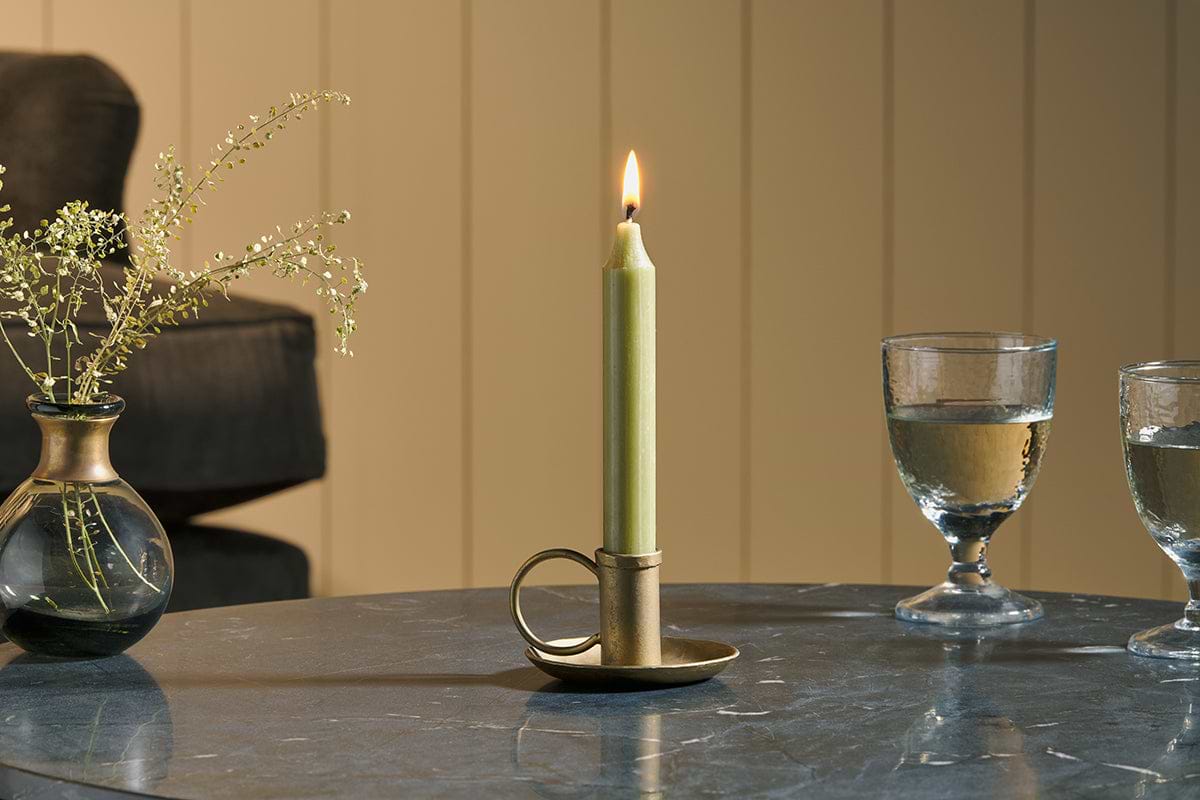 An Amri Iron Candlestick by nkuku in antique brass holds a lit green dinner candle on a round marble table, accompanied by two glasses of water and a small vase of dried flowers. In the background are a dark sofa and light vertical paneling.