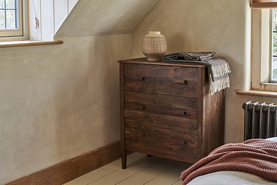 The nkuku Anbu Acacia Chest of Drawers in Washed Walnut stands in the corner, topped with a beige textured vase and folded gray throw. Sunlight from nearby windows brightens the neutral-toned bedroom storage space.
