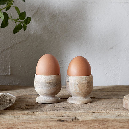Two brown eggs in nkukus Awa Wooden Egg Cup - Natural (Set of 2) sit on a rustic breakfast table with tableware, bread, a potted plant, and a cloth against a light textured wall.