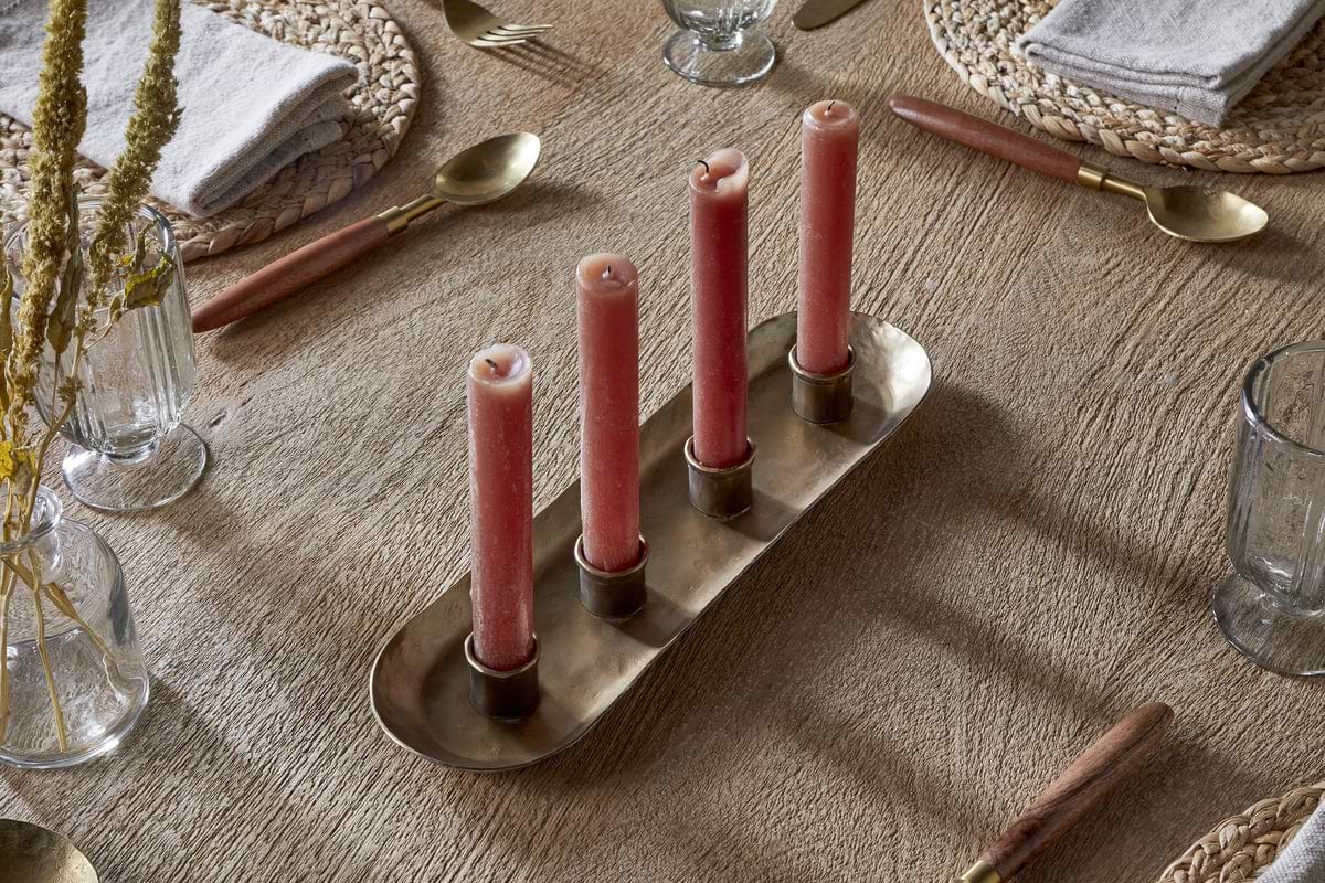 A wooden dining table set for a meal features woven placemats, gold utensils with wooden handles, clear glasses, gray napkins, and the nkuku Amita Long Candle Holder in antique brass as a centrepiece holding four pink candles. Vases with dried grasses complete the scene.