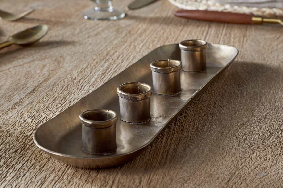 The nkuku Amita Long Candle Holder in antique brass features a rectangular design with rounded corners and four small cylindrical cups in a row, creating an elegant centrepiece for your dining table.