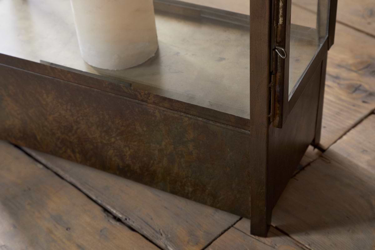 Close-up of the Akoko Wide Lantern by nkuku, featuring a brown patina finish on glass and metal, with a candle inside, placed on a rustic wooden floor.