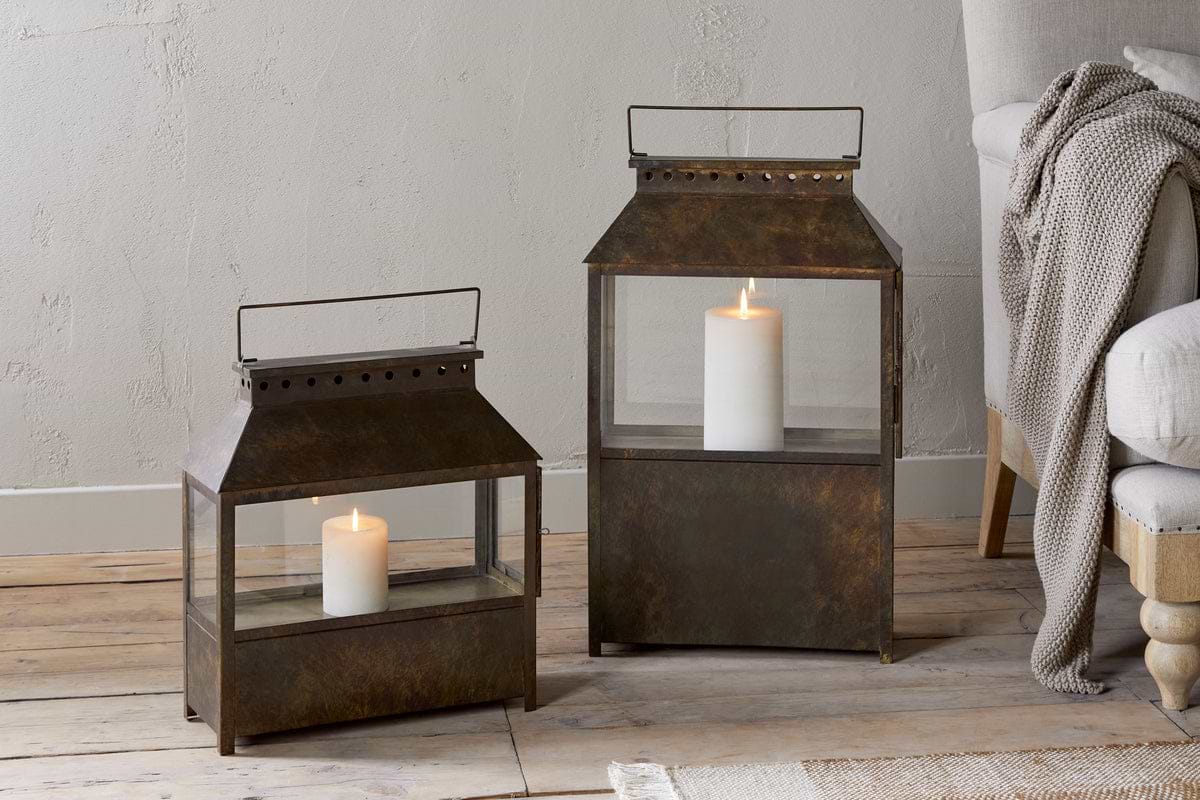 Two square Akoko Wide Lanterns by nkuku, finished in brown patina and each holding a lit white pillar candle, rest on a wooden floor. A beige armchair with wooden legs and a knitted throw sits against a textured neutral wall.