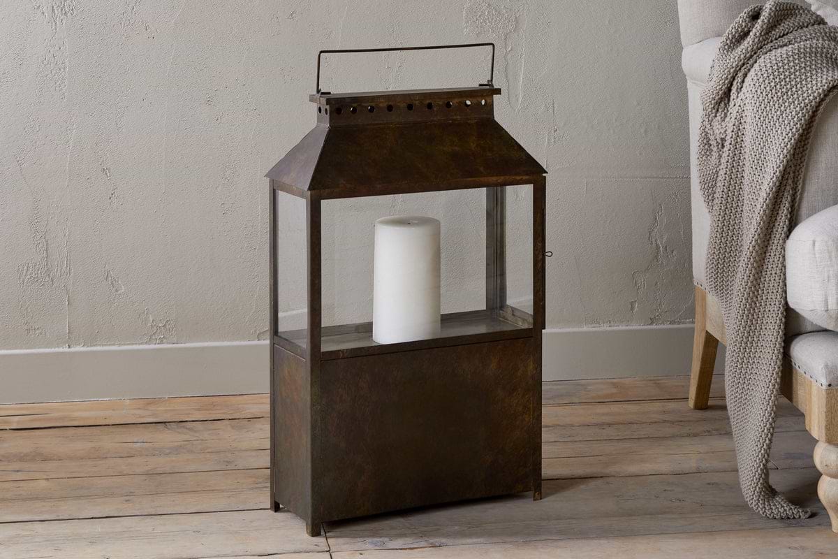 The nkuku Akoko Wide Lantern in Brown Patina with glass panels and a white pillar candle adds cozy charm beside a light-upholstered chair layered with a knit blanket.