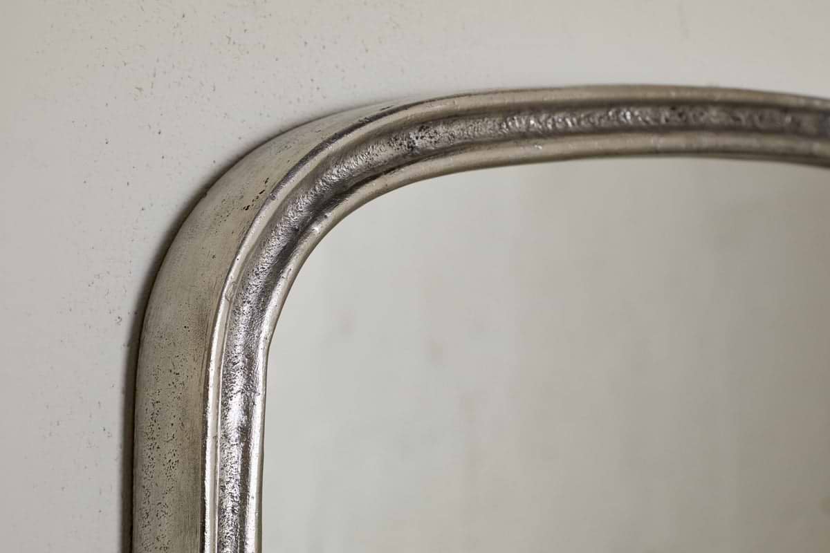 Close-up of the top left corner of the nkuku Almora Wall Mirror - Antique Nickel, featuring a textured metallic aluminium frame against a light-colored wall.