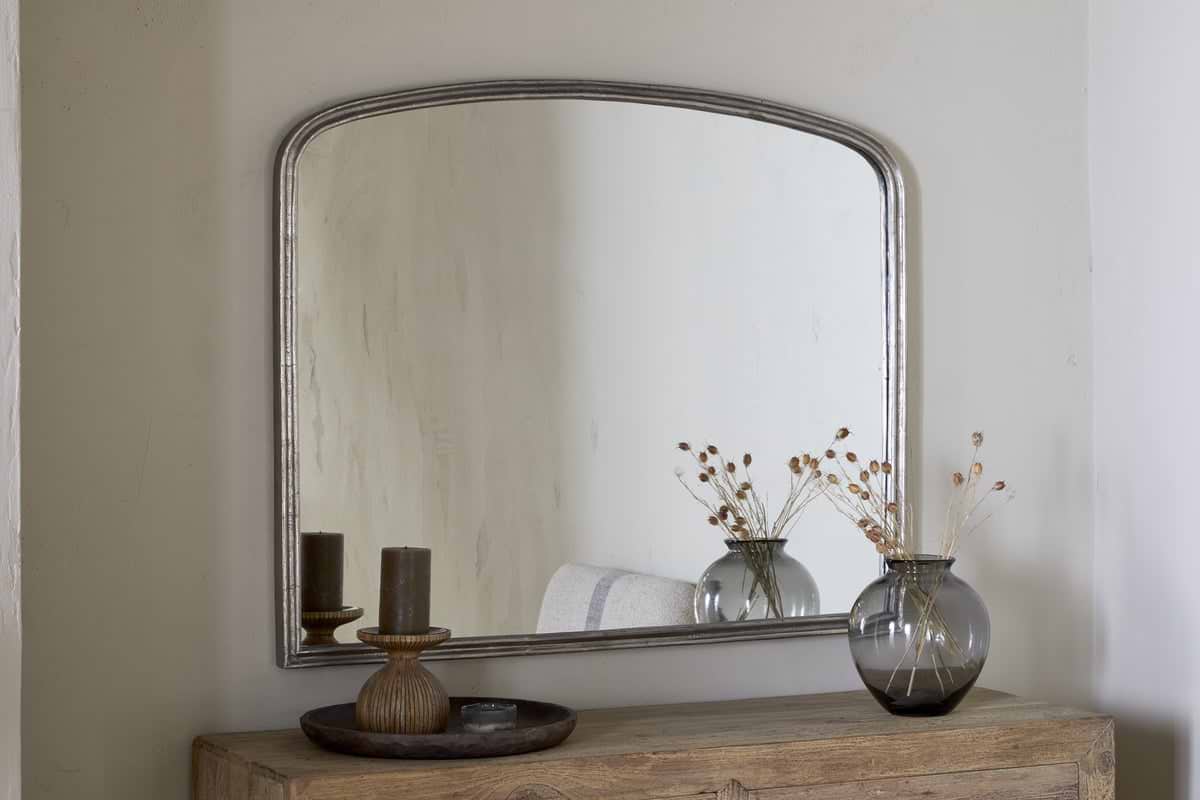 The nkuku Almora Wall Mirror in Antique Nickel, with its arched aluminium frame, hangs on a light wall above a rustic wooden table. A gray glass vase with dried flowers and a wooden tray with two candles decorate the table.