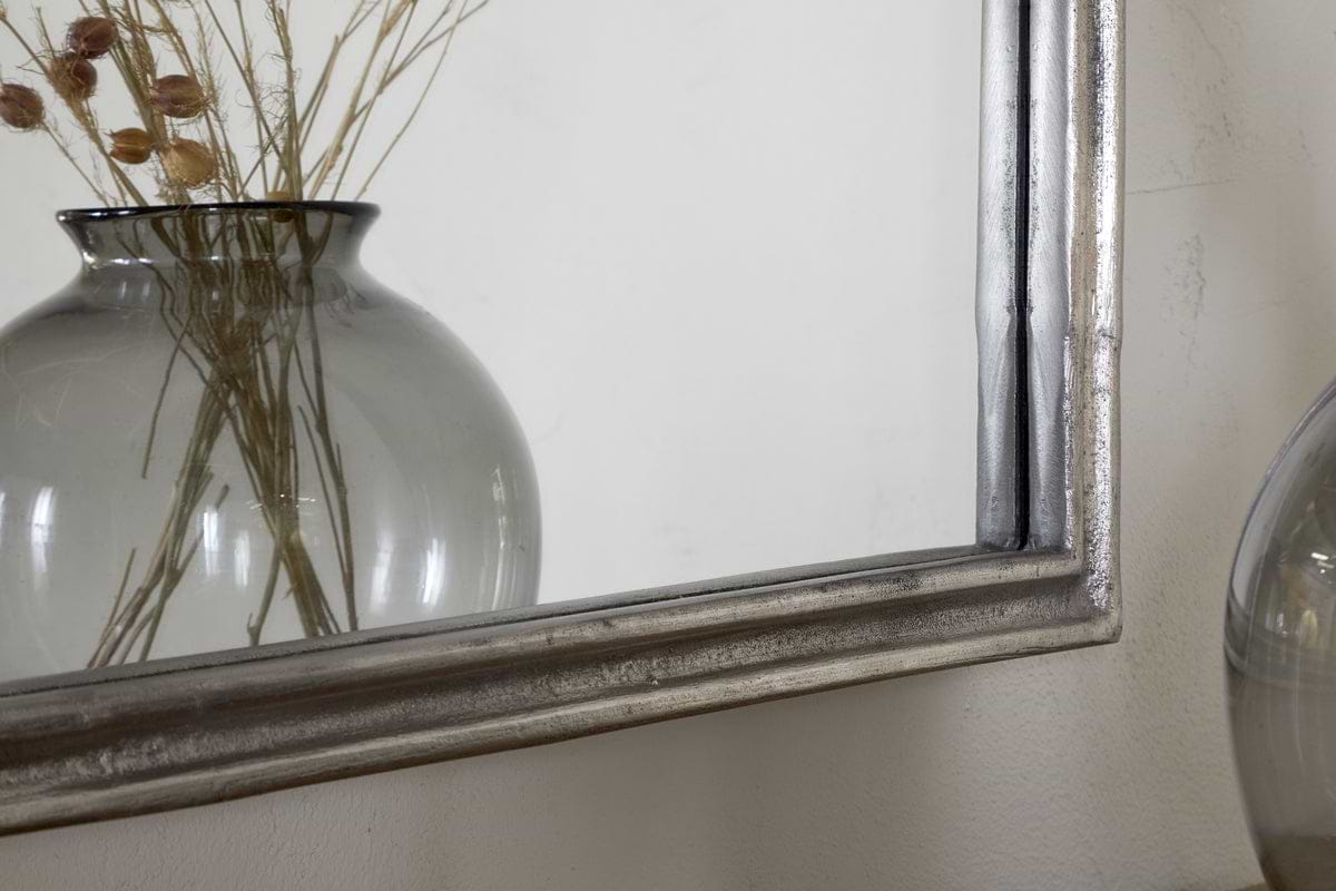 A close-up of dried plants in a gray glass vase, partially reflected in the nkuku Almora Wall Mirror (Antique Nickle) against a white wall.