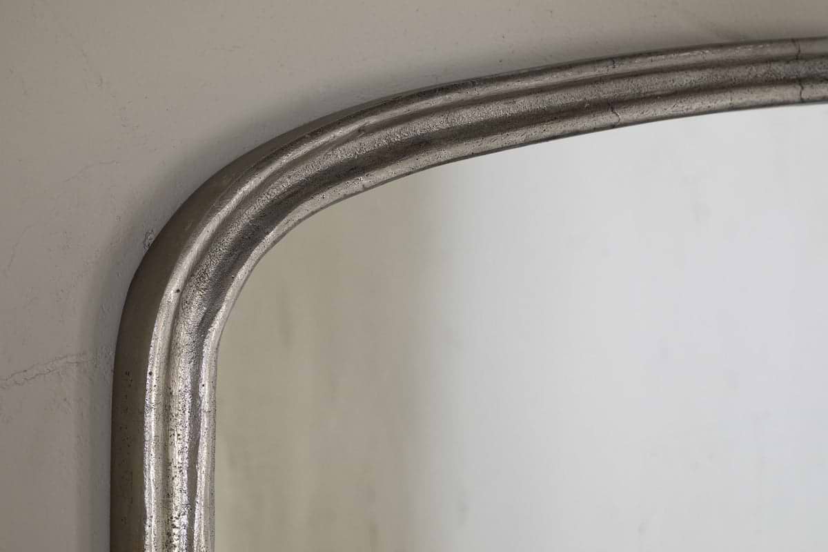 Close-up of the top left corner of the nkuku Almora Wall Mirror - Antique Nickle, featuring a rectangular design with rounded edges and a metallic, slightly textured aluminium frame against a plain, light-colored wall.