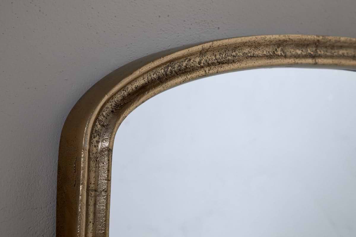 Close-up of the top corner of the nkuku Almora Wall Mirror, Extra Small, in antique brass with a distressed finish, set against a light gray wall.