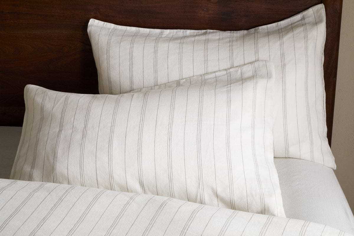 Two white pillows with thin, vertical gray stripes rest against a dark wooden headboard. Pair them with nkuku’s Anta Hemp & Cotton Housewife Pillowcases in Sage Green for a fresh touch. A matching striped blanket is partially visible below.