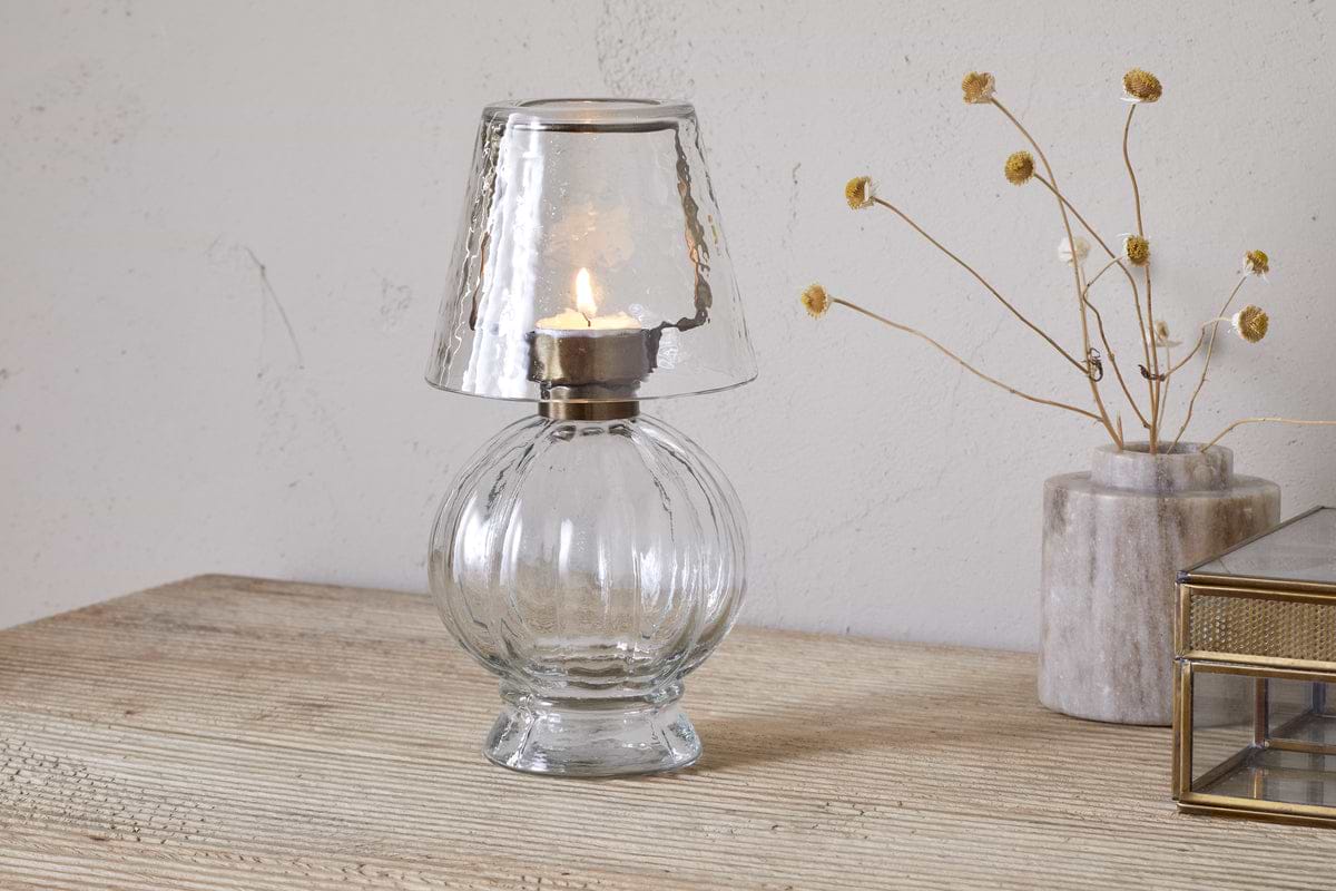 The nkuku Adesola Glass Tealight Holder Lamp in clear sits on a wooden surface beside tealight holders, a marble vase with dried flowers, and a gold-edged glass box, all set against a light-colored wall.