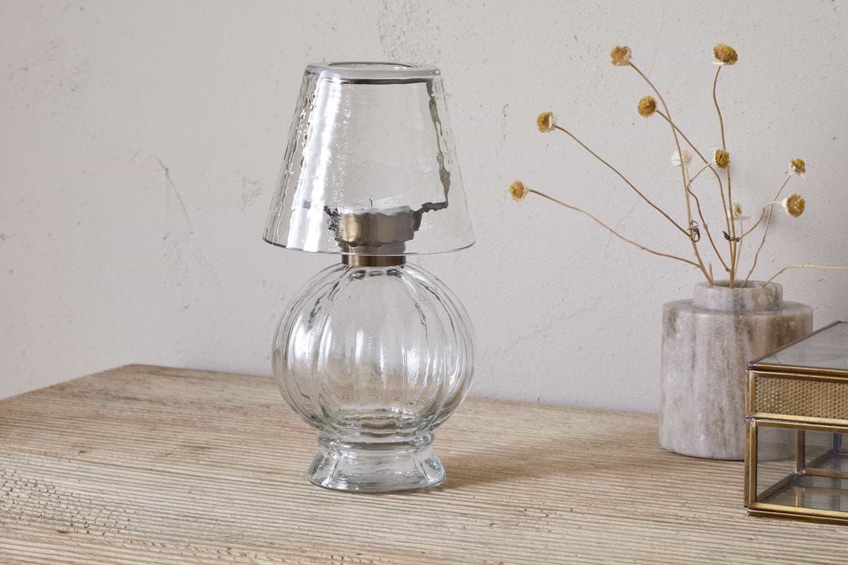The nkuku Adesola Glass Tealight Holder Lamp in clear glass sits on a wooden surface beside dried yellow flowers and a glass box, set against a light, textured wall.