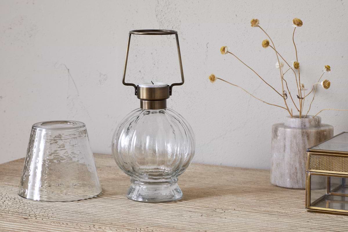The nkuku Adesola Glass Tealight Holder Lamp in clear, featuring a brass handle, rests on wood alongside an upside-down glass, a marble vase with dried flowers, and a glass box before a textured light wall.