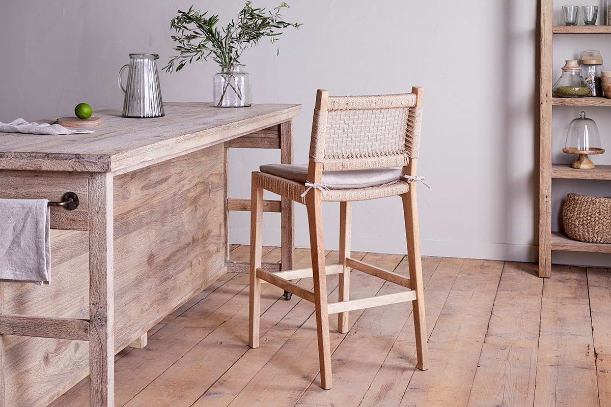 An Aamani Mango Wood & Cord Counter Chair by nkuku sits at a rustic kitchen island topped with a glass jug, lime, and a small vase of greenery. Shelves in the background display jars, baskets, and dishes for added charm.