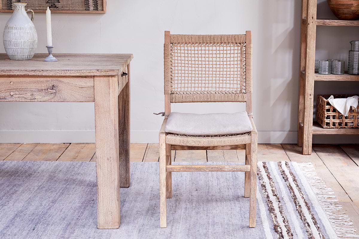 The nkuku Aamani Mango Wood & Cord Dining Chair – Natural, featuring a woven recycled paper cord back and linen seat cushion, sits by a reclaimed wood table with a white vase and candle; wooden shelves and striped rug complete the scene.
