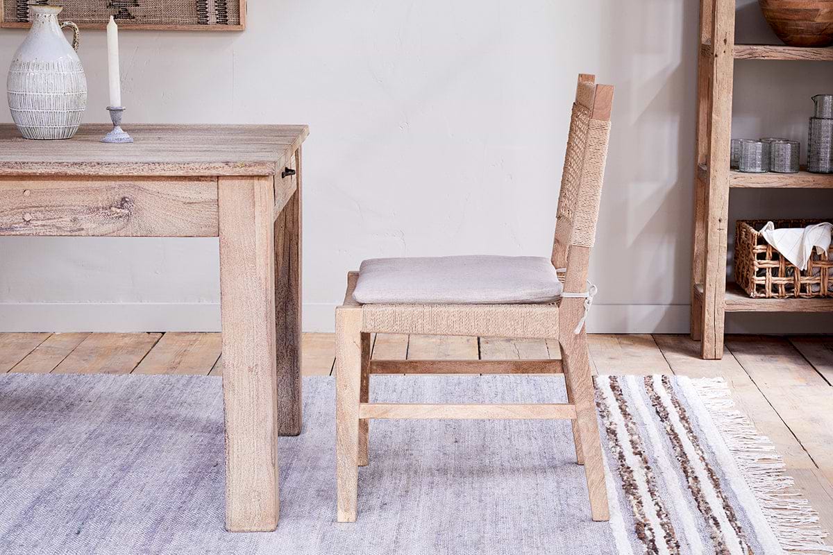 The Aamani Mango Wood & Cord Dining Chair by nkuku, crafted from natural mango wood with a beige cushion, pairs with a matching table. A woven rug, shelf of arranged dishes, and soft lighting complete the rustic, neutral-toned space.