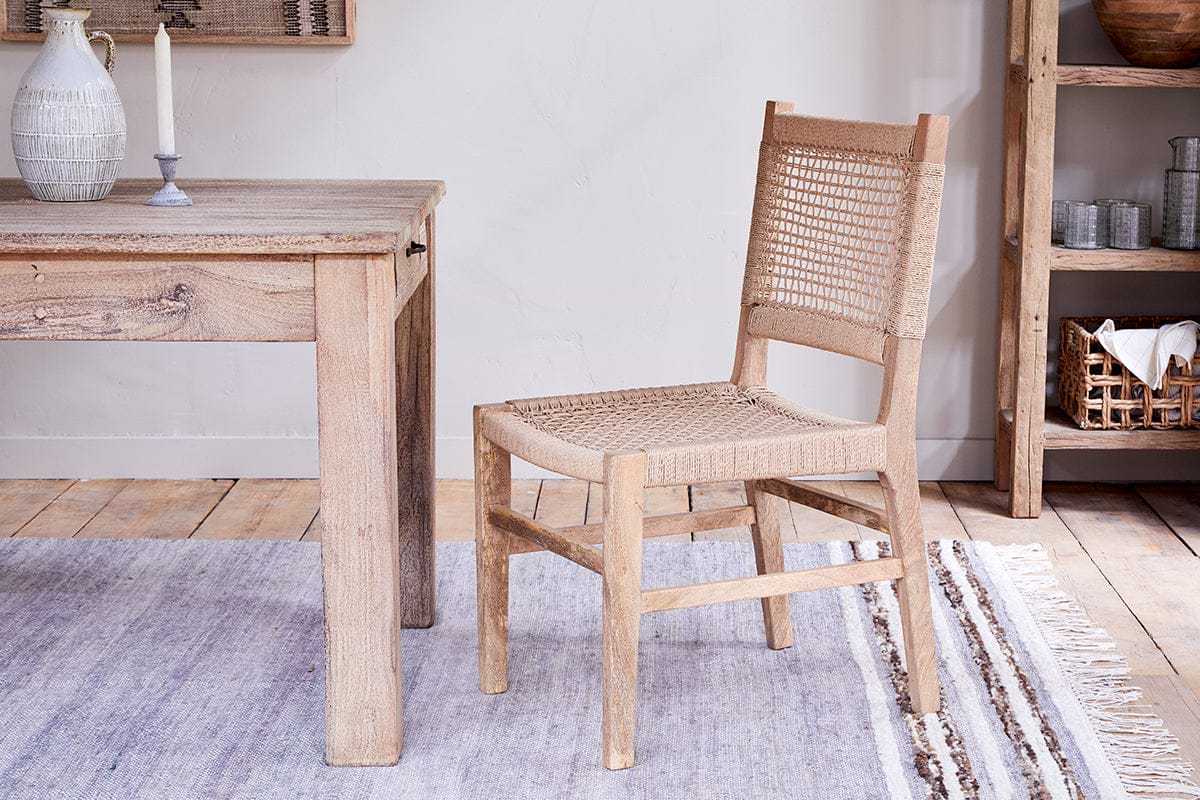 The nkuku Aamani Mango Wood & Cord Dining Chair in Natural sits by a matching table on a light gray rug atop wooden floors, with decor-filled shelves adding to the cozy, natural ambiance.