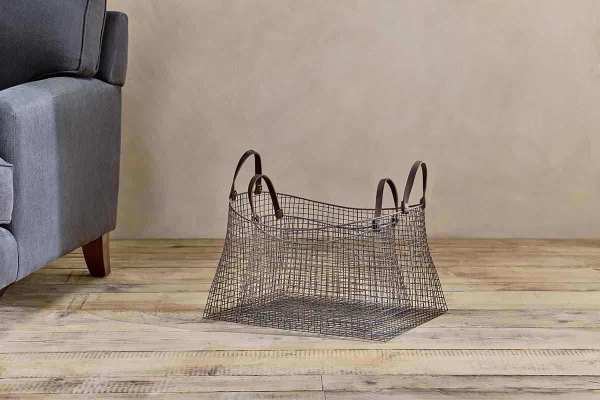 The Adah Iron Baskets - Brown (Set of 2) by nkuku, featuring leather handles and a geometric design, sit beside a gray sofa on a light wood floor, complementing the neutral, minimalist decor against the beige wall.