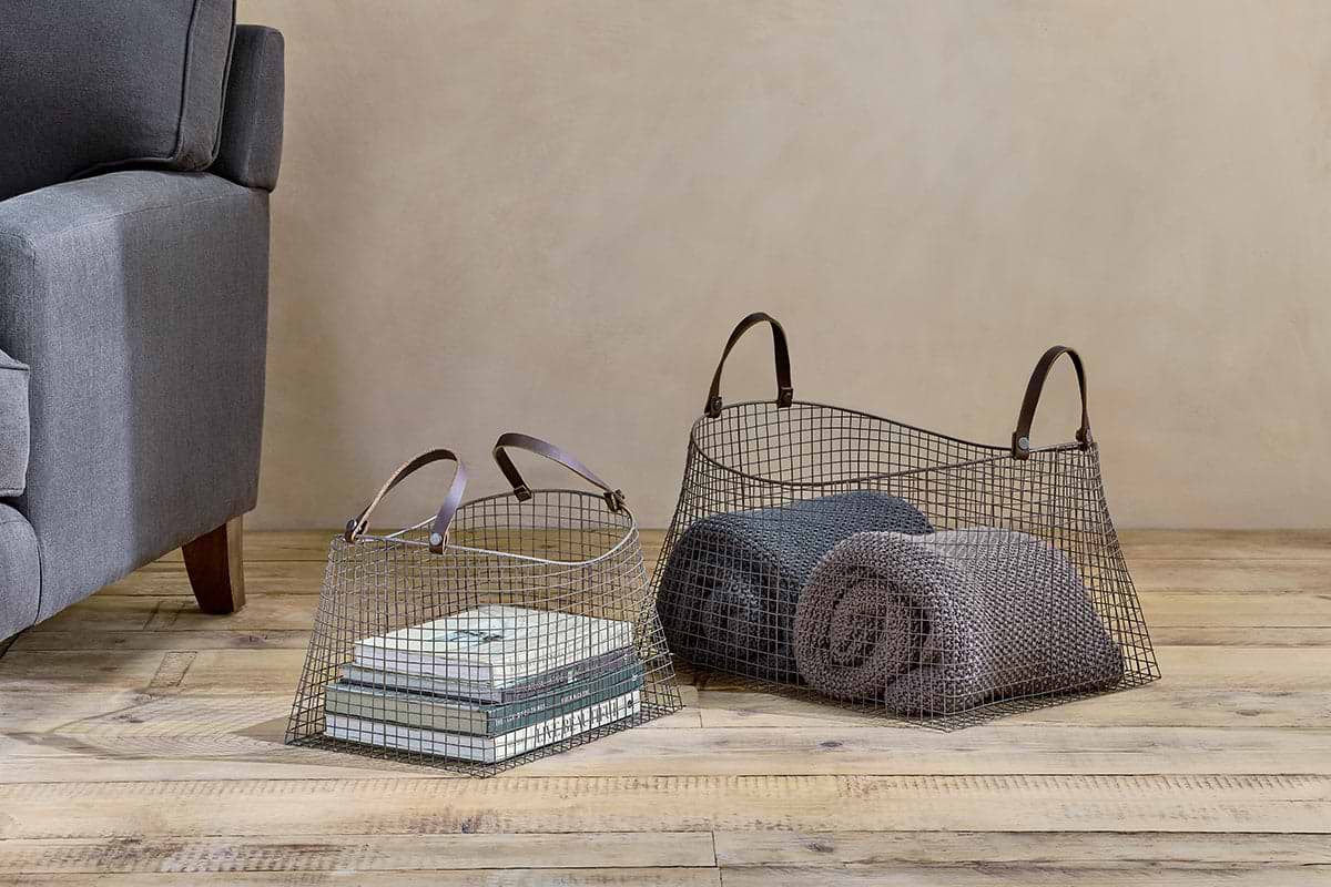 Two Adah Iron Baskets - Brown by nkuku rest on a wooden floor beside a gray couch. One basket stores books, while the other holds two rolled blankets in gray and taupe, set against a beige wall.