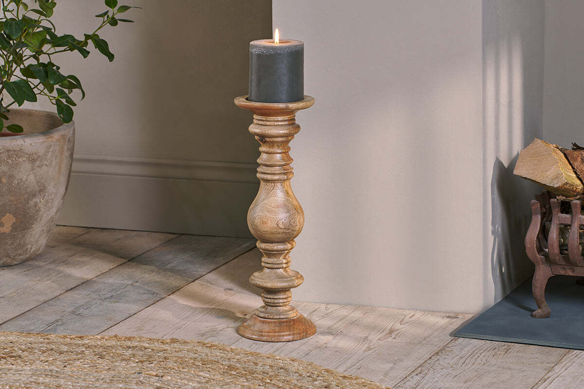 The Akello Candle Stick by nkuku, featuring a dark gray pillar candle, sits on a light wood floor near a woven rug, potted plant, and metal log holder—creating cozy fireplace decor as the candle casts a warm glow.