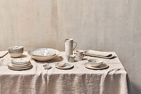 A beige tablecloth covers a table set with ceramic dishes, bowls, and nkuku’s Ama Short Splatter Mug (Set of 2), plus a cream-colored splatter pitcher. The plain light wall highlights the rustic, minimalist vibe.