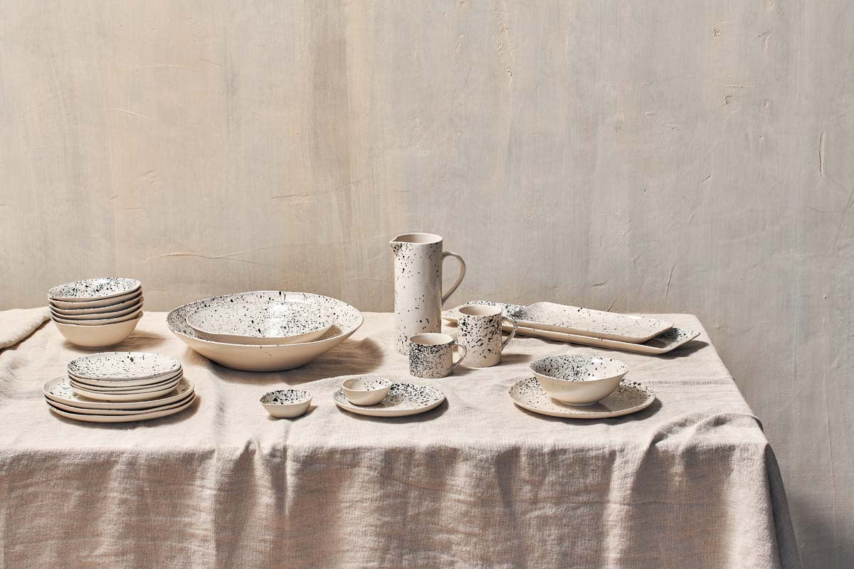 A beige tablecloth covers a table set with ceramic dishes, bowls, and nkuku’s Ama Short Splatter Mug (Set of 2), plus a cream-colored splatter pitcher. The plain light wall highlights the rustic, minimalist vibe.