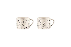 The Ama Short Splatter Mug Set by nkuku features two white handmade mugs with black splatters, rounded bodies, and large handles, displayed side by side on a plain white background in a minimalist, well-lit photo.