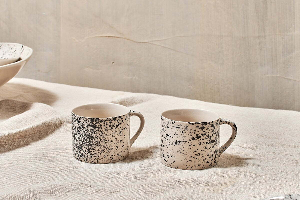 Two nkuku Ama Short Splatter Mugs (Set of 2) in beige ceramic with black splatter glaze and simple handles are displayed on a light-colored textured fabric.
