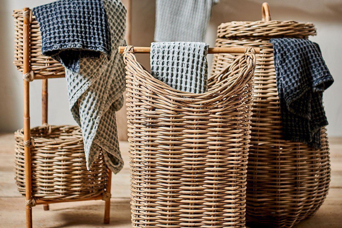 Three Amandi Rattan Laundry Bins by nkuku in different sizes are arranged on a wooden floor, two filled with blue and gray towels. A matching nkuku rattan stand with a towel completes the cozy, rustic bathroom storage setup.