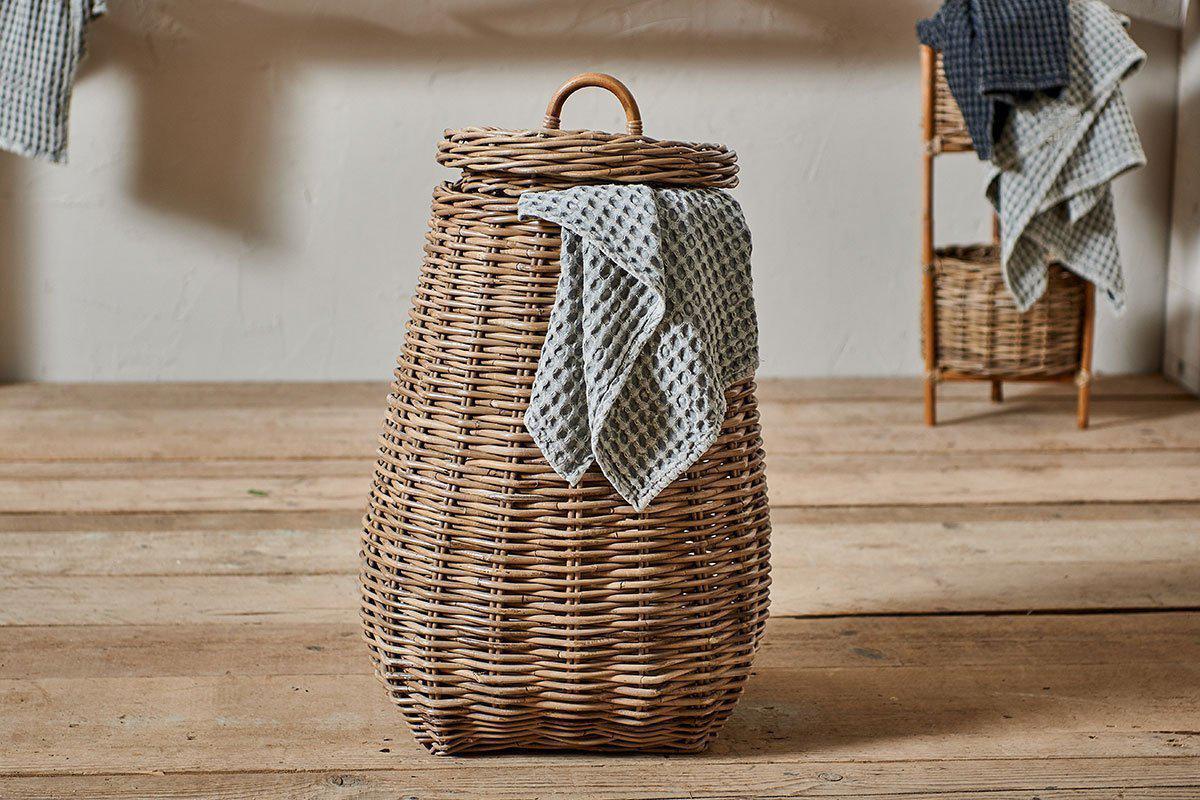 The Amandi Rattan Laundry Bin by nkuku stands on a wooden floor with a blue and white checkered cloth draped over its side. In the background are stacked towels and a smaller natural rattan basket against a plain wall.