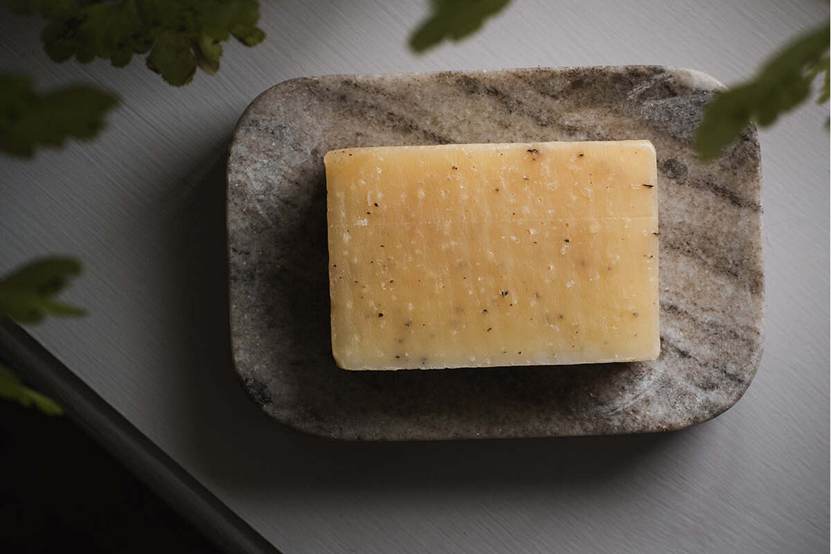 A rectangular, yellowish soap with small specks sits on the nkuku Asana Marble Soap Dish - Grey, its smooth oval surface highlighted by soft light and framed by green leaves.
