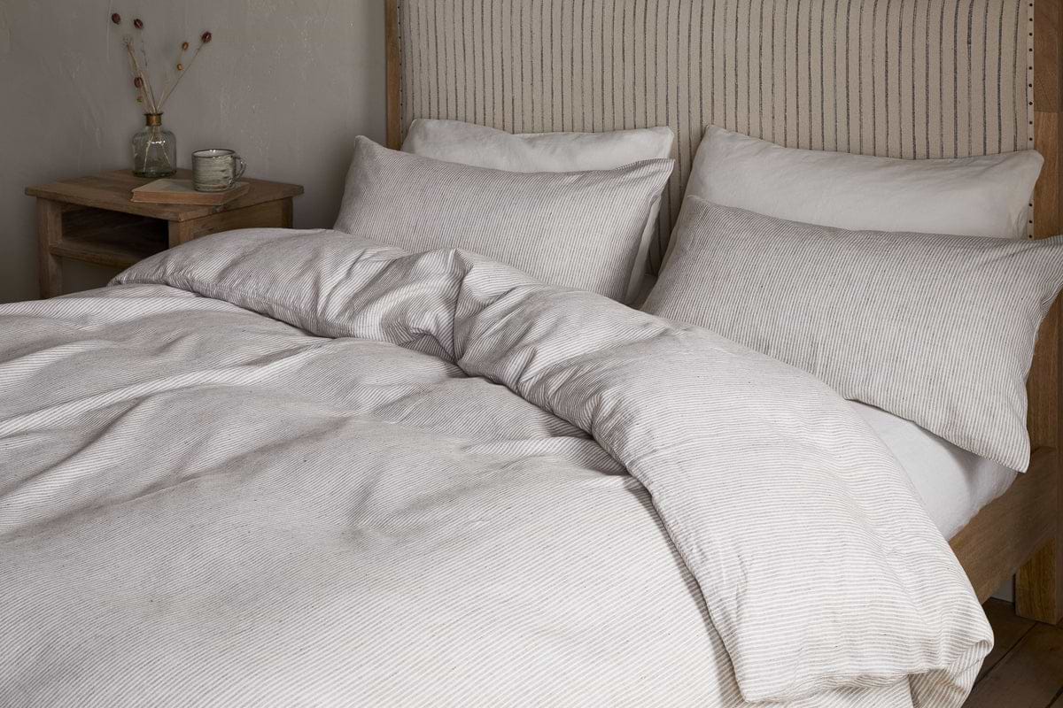 A neatly made bed features the nkuku Borba Hemp & Cotton Duvet Cover in Espresso, with matching pillows, beside a wooden nightstand topped with a clear vase of dried flowers and a small glass jar.