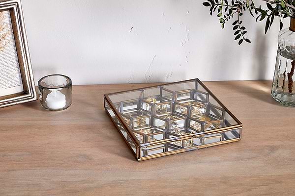 The nkuku Bequai Honeycomb Box, a square glass piece with gold trim, rests on wood holding elegant jewelry. Nearby sit a glass votive candle, a photo frame, and a vase with green foliage for stylish jewellery storage.