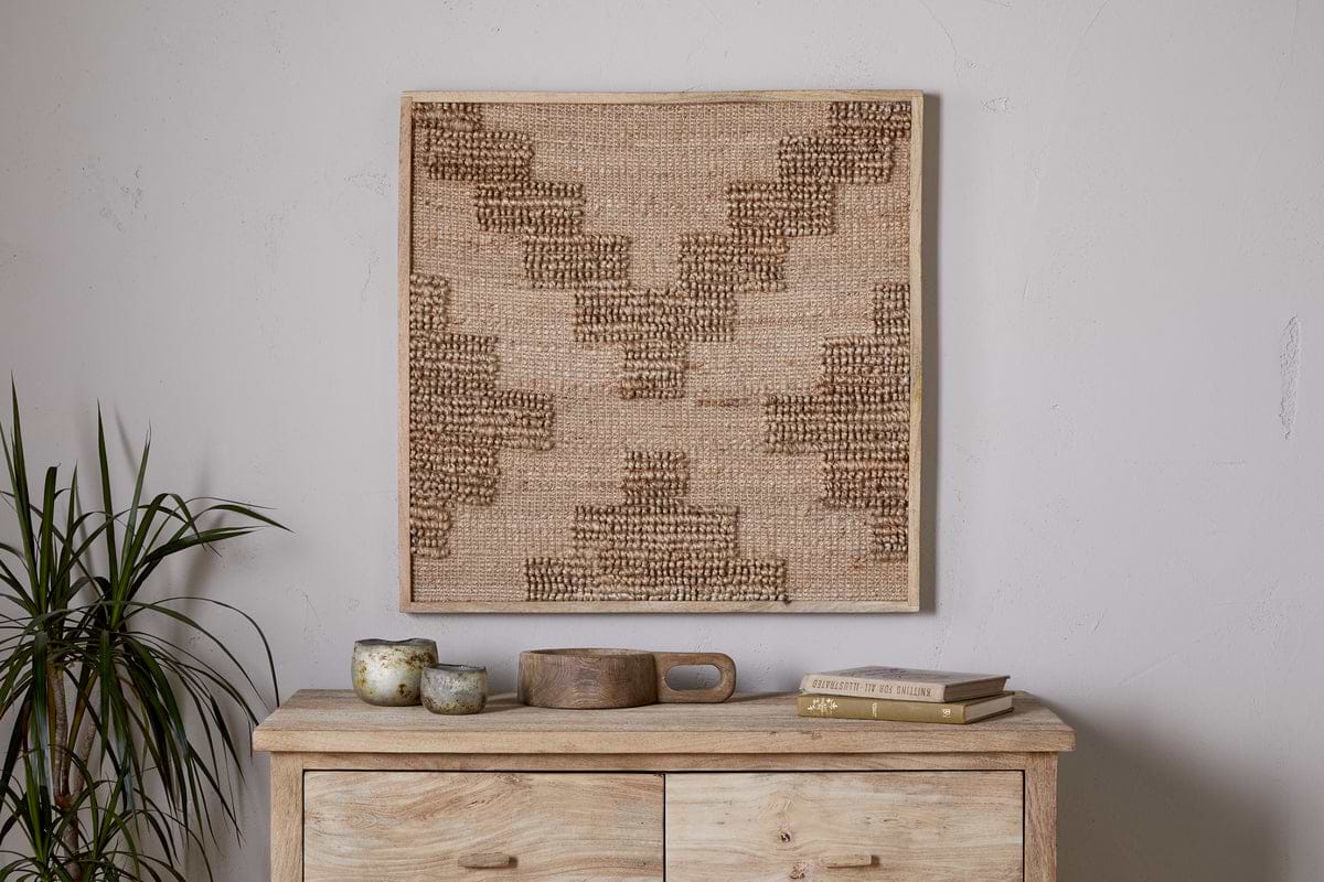 A light wooden chest holds small rustic cups, a tray, and books. Above, the nkuku Bawal Wall Art - Cross - Natural with tan and brown geometric patterns hangs on a white wall. To the left stands a leafy green plant.