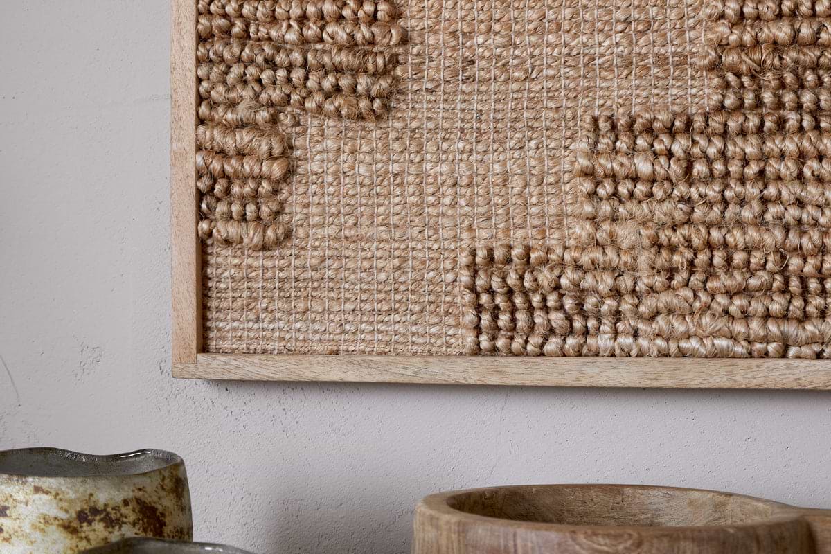 A close-up of nkuku's Bawal Wall Art - Cross - Natural features thick tan fibers in a sustainable mango wood frame with a textured geometric pattern. In front, a rustic ceramic vase and wooden bowl sit on a light gray surface.