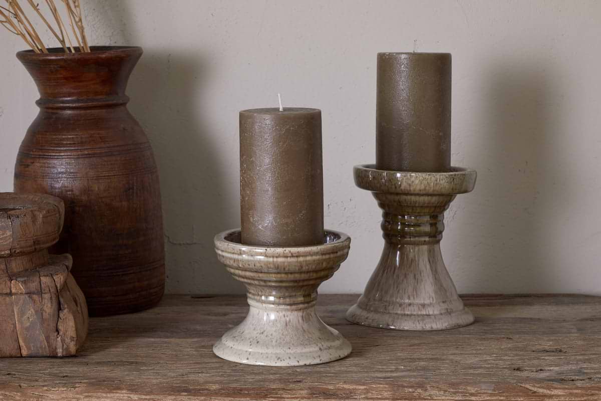 Two brown pillar candles sit in nkuku Cabra Reactive Glaze Candle Holders in Olive & Natural on a wooden surface, with a tall brown clay vase of dried grass and stacked wooden boxes creating an earthy, rustic vintage decor.