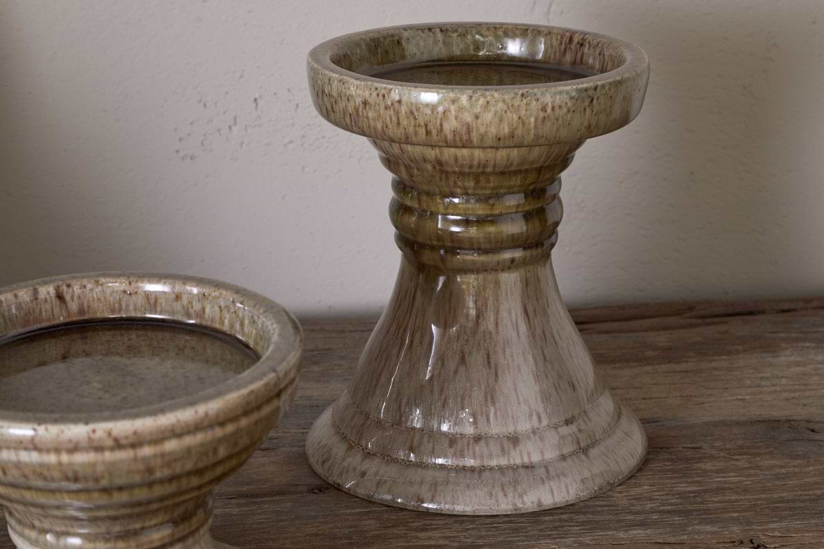 Two Cabra Reactive Glaze Candle Holders by nkuku, in olive and natural tones with a glossy, speckled finish, sit on a rustic wooden surface. One holder is fully visible with its flared base, while the other appears partially in the corner.