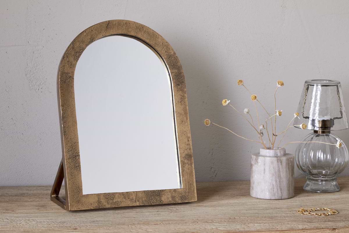 The nkuku Cayce Cast Aluminum Vanity Mirror in Antique Brass sits on a wooden dressing table beside a marble vase with dried flowers, a clear glass oil lamp, and a gold bracelet against a light, textured wall.