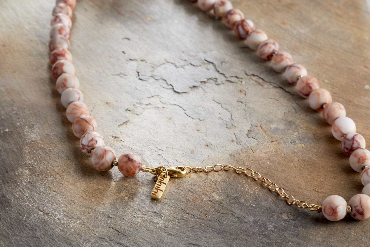 The Cerita Necklace - Rhodochrosite by nkuku features marbled pink and white semi-precious stones and a gold chain extender, displayed on an earthy textured surface—a versatile accessory for any occasion.