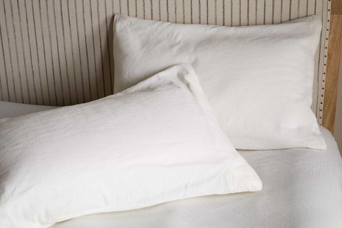 Two Cela Hemp & Cotton Housewife Pillowcases by nkuku in natural shade rest on a light bed with a striped headboard. One pillow stands upright, the other lies flat, suiting different bedroom styles with their breathable hemp and cotton blend.