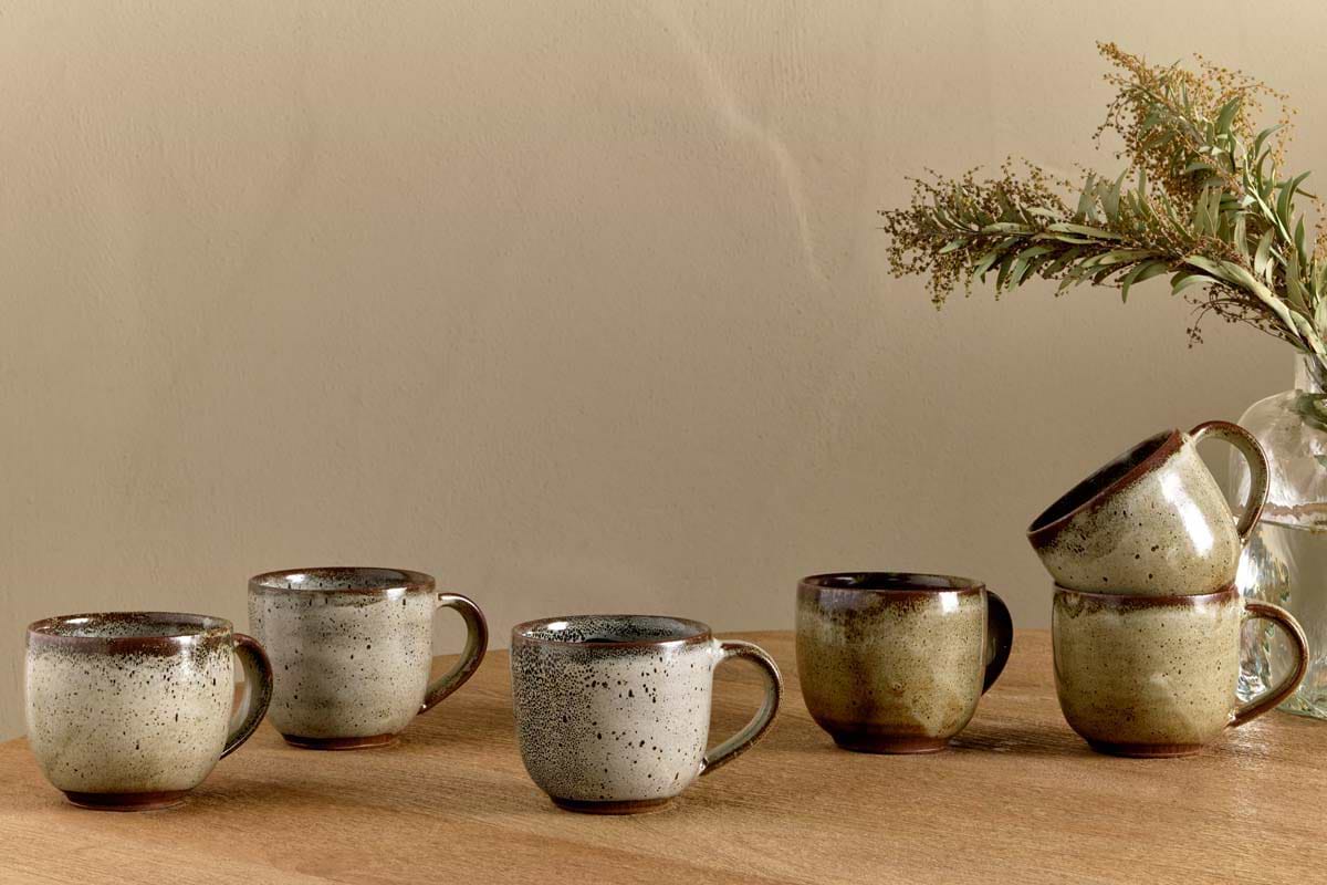 Five nkuku Cam Coffee Mugs - Mocha (Set of 2) with reactive glaze are arranged on a wooden surface; three stand alone, two are stacked. A glass vase with dried greenery is set in the background against a beige wall.