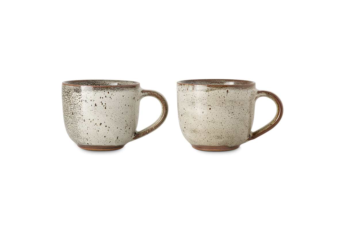 The nkuku Cam Coffee Mug - Mocha (Set of 2) features rustic mugs with handles, reactive speckled beige glaze, brownish rims, textured patterns, slightly rounded shapes, and warm brown unglazed bases displayed side by side on white.