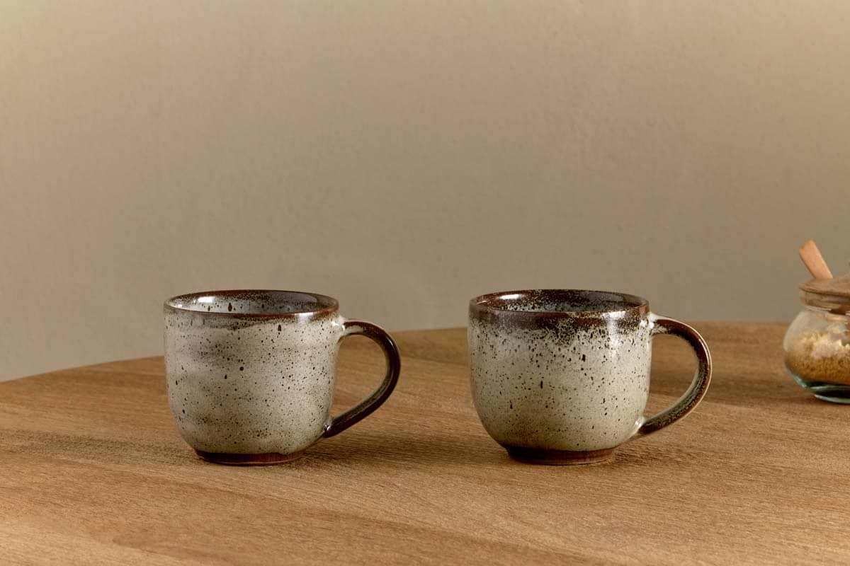 Two Cam Coffee Mugs - Mocha by nkuku (set of 2) with a speckled reactive glaze rest on a wooden table. A glass jar with a wooden lid appears on the right, all set against a neutral beige wall backdrop.