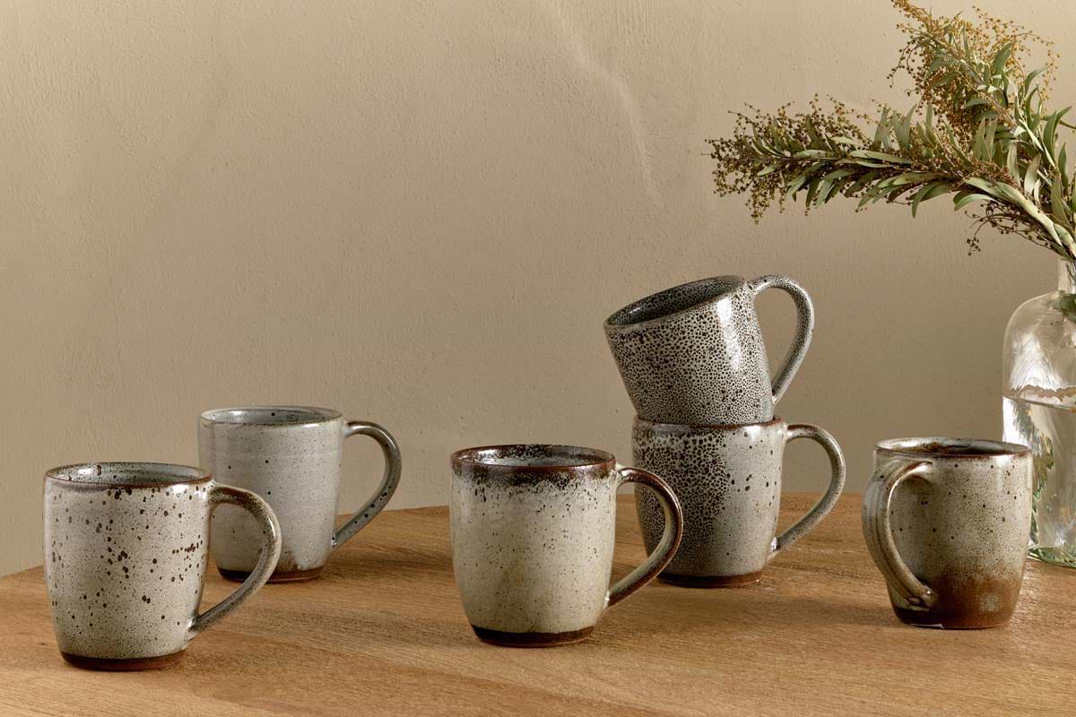 Six nkuku Cam Large Mugs in mocha, sold as a set of 2, feature earthy tones and reactive glaze. Arranged on wood with two mugs stacked, they sit beside a glass vase of eucalyptus against a neutral beige wall.