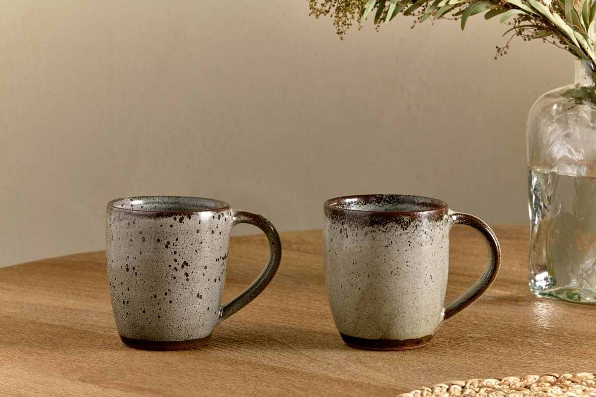 Two nkuku Cam Large Mug - Mocha (Set of 2) with brown rims and speckled gray glaze rest side by side on a wooden table. A glass vase with water and green foliage is partially visible in the background.
