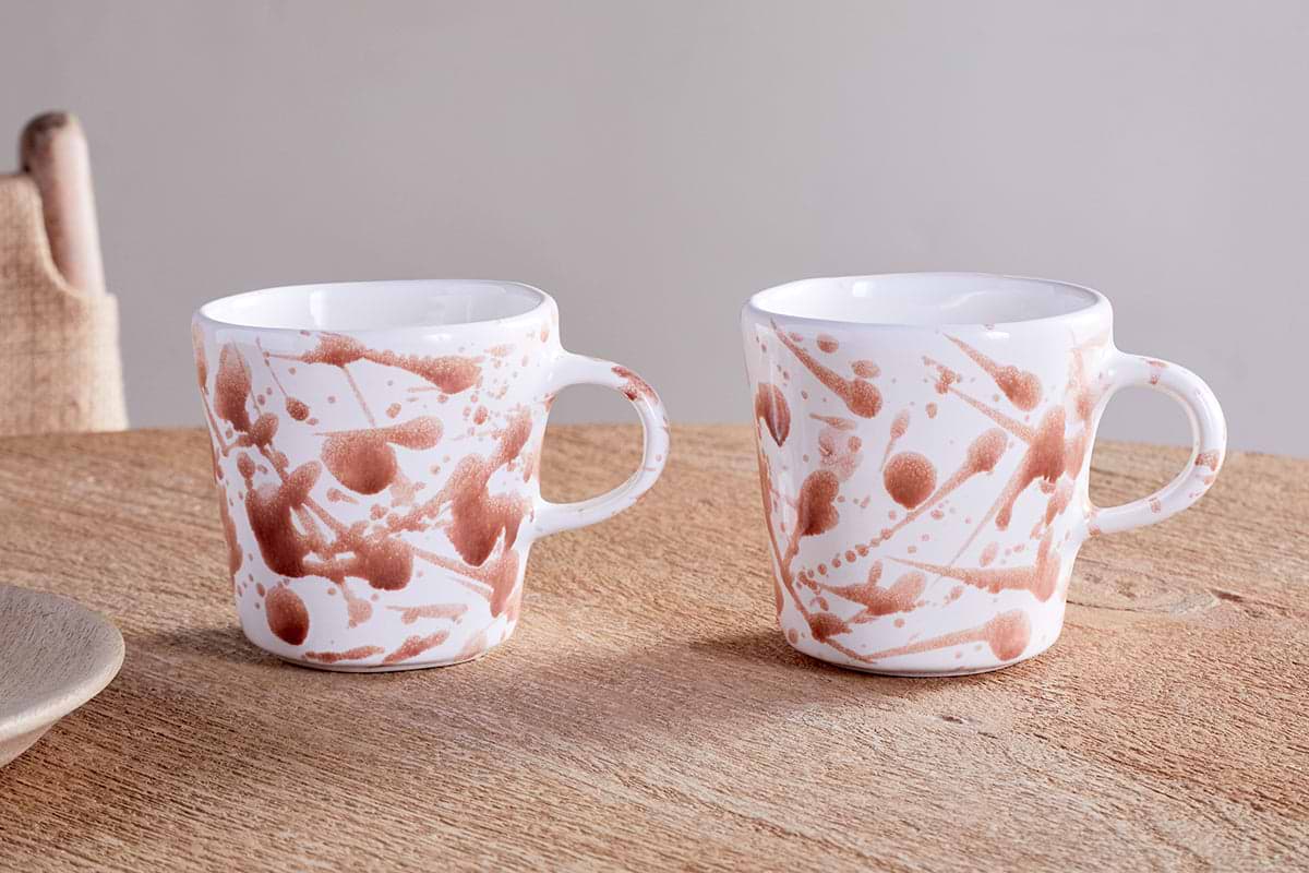 Two nkuku Canela Mug - Dusty Terracotta (Set of 2) feature white glaze with rust-hued splatter, round handles, and a tapered shape. They sit on a wooden table with linen napkins in the background.