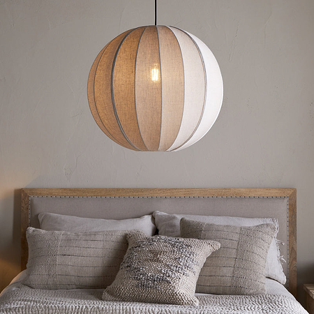 A neutral-toned bedroom showcases a bed with pale bedding, textured pillows, a wooden headboard, a small wooden nightstand with a ceramic bowl, and the nkuku Dharini Linen Lampshade - Natural hanging above.