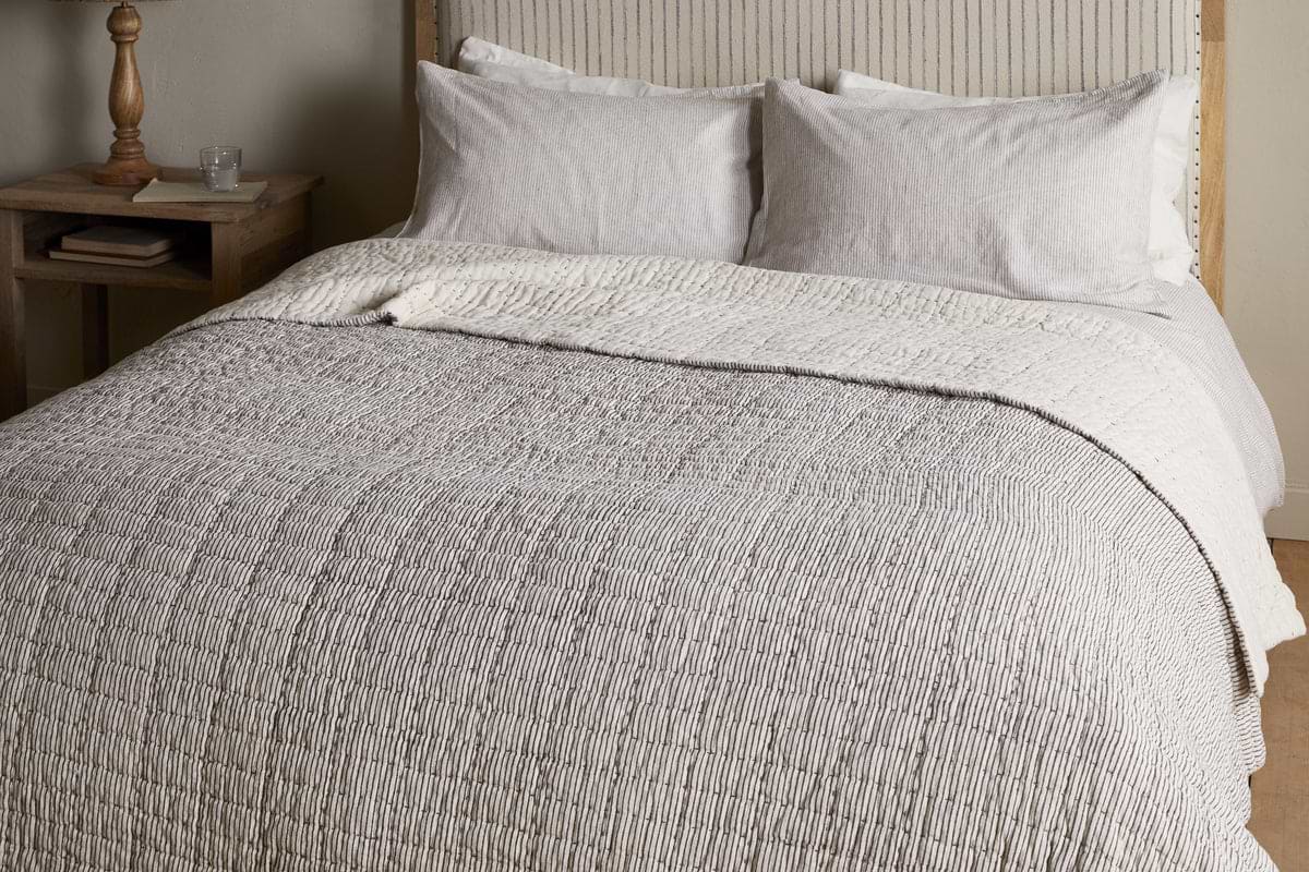The nkuku Dipti Cotton & Linen Block Print Bed Quilt in Charcoal adds comfort and style, featuring a soft cotton linen blend with a subtle charcoal stripe design—perfect for cozying up in bed.