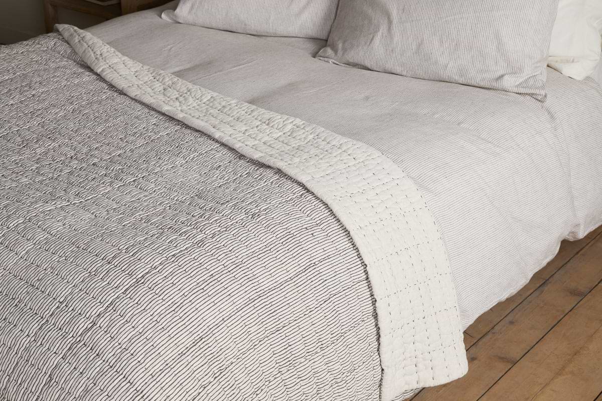 A neatly made bed features the nkuku Dipti Cotton & Linen Block Print Bed Quilt in Charcoal. Its subtle block print and cozy cotton-linen blend bring a minimalist, textured look that complements the room’s wooden floor.