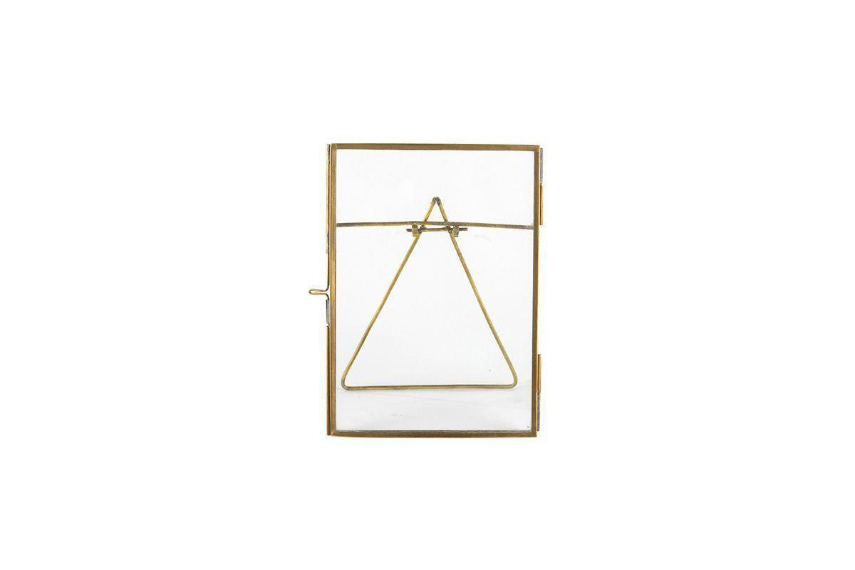 The nkuku Danta Antique Brass Frame features an upright rectangular glass design with a slim brass border. Hand-forged metal wire forms a centered triangle attached to the top corners, set against a plain white background.
